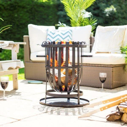 A cozy outdoor patio showcases the La Hacienda Vancouver Firebasket at its center, encircled by wicker seating adorned with white cushions. Skewers laden with food sizzle above the flames, capturing the essence of outdoor cooking. Sunlight bathes the setting, while two glasses of wine rest gracefully on the ground.