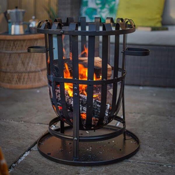 The La Hacienda Vancouver Firebasket, crafted by La Hacienda, features vertical bars that securely hold burning logs while casting a warm glow. It is placed on a stone patio surrounded by wicker furniture adorned with colorful cushions, creating a cozy outdoor haven ideal for gatherings or outdoor cooking.