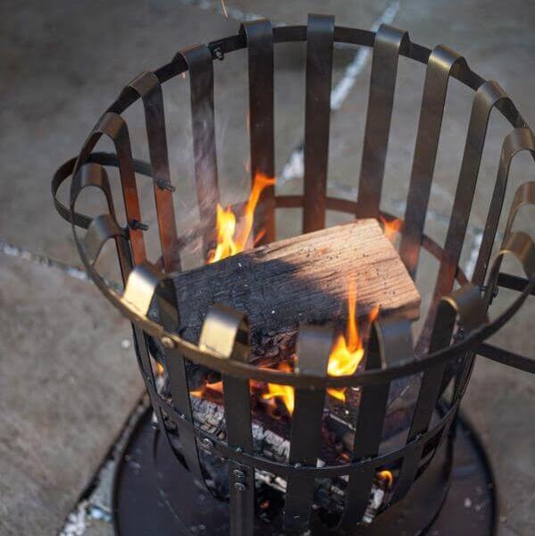 The La Hacienda Vancouver Firebasket, crafted by La Hacienda, features a wooden log burning inside and produces vibrant orange flames. Ideal for outdoor cooking, this fire basket is set on a concrete surface with some small ashes dispersed around it.