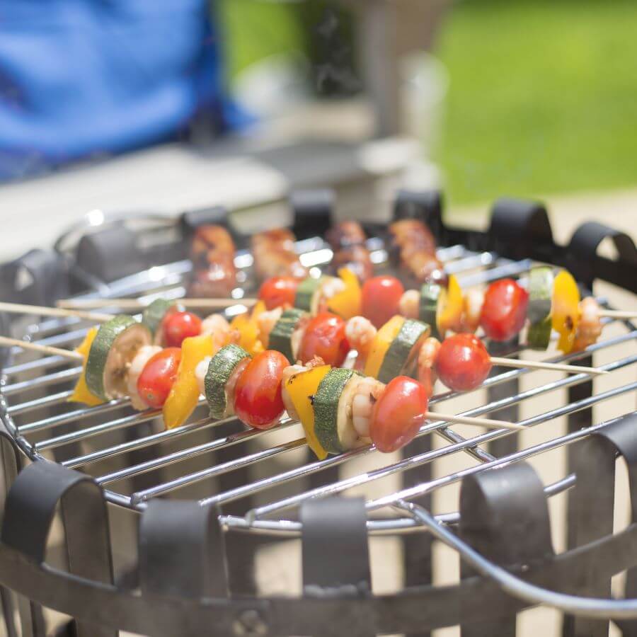 Vegetable skewers with cherry tomatoes, zucchini, mushrooms, and yellow bell peppers sizzle over the La Hacienda Vancouver Firebasket, showcasing the art of outdoor cooking. In the background, a few sausages lie basking in the sunshine.