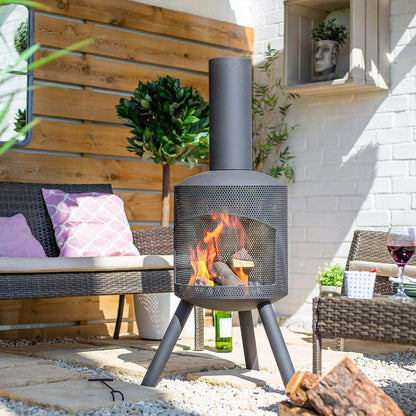 The La Hacienda Santana Fireplace, known for its modern design and robust steel construction, burns logs in its metal chimney. It is placed on a stone patio adorned with wicker furniture and purple cushions. Close by are a glass of red wine and a potted plant, all surrounded by a wooden fence and a white brick wall.