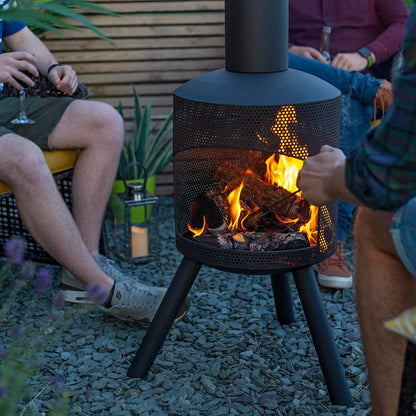 A group of people gathers around a La Hacienda Santana Fireplace on a gravel surface, with its vibrant flames flickering within the modern design. Encircled by diverse plants and a wooden panel backdrop, one person attends to the fire while others unwind, drinks in hand, in this tranquil environment.