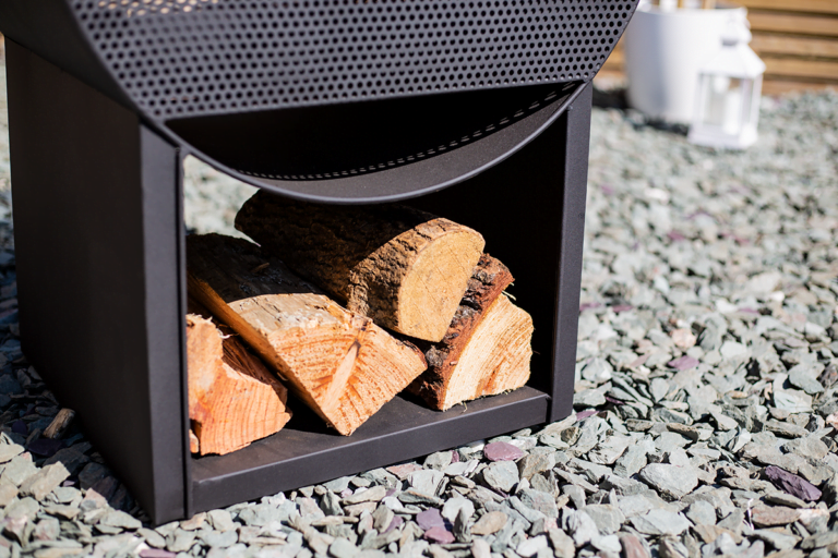 A contemporary black metal log holder crafted from durable steel sits on a bed of gray stones with neatly stacked firewood, basking in the sunlight that accentuates the wood's texture and grain. A white lantern complements this La Hacienda Camacha Fireplace setting.