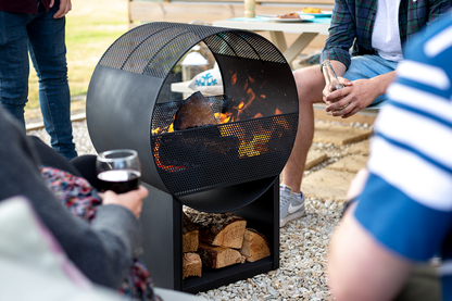 A group of people gathered around the La Hacienda Camacha Fireplace, situated on a gravel surface. Its durable steel construction and convenient log storage below provided a warm ambiance, as one individual held a drink, creating a relaxed social setting.