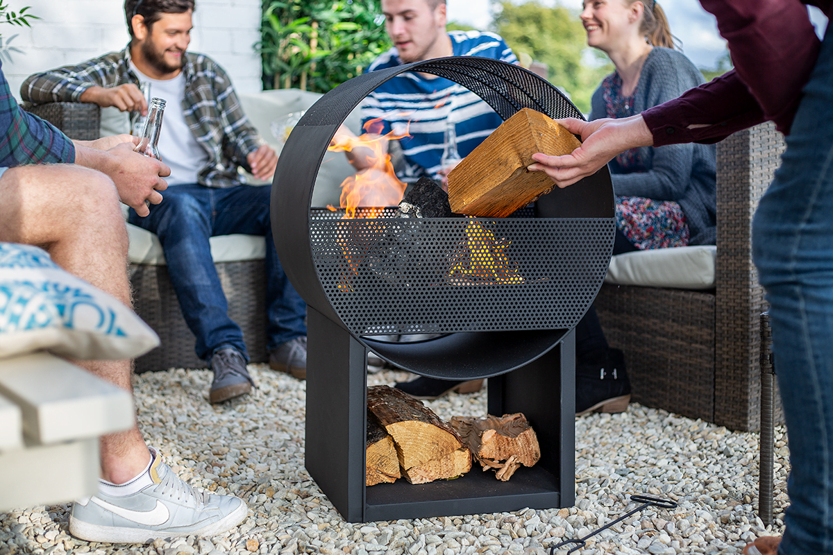 A group of people gathered around the contemporary La Hacienda Camacha Fireplace, featuring a sleek circular design; one person is adding a log to the flames. Others are seated on patio furniture, sipping drinks and relishing the cozy ambiance of the gravel-covered patio, which is complemented by its sturdy steel construction.