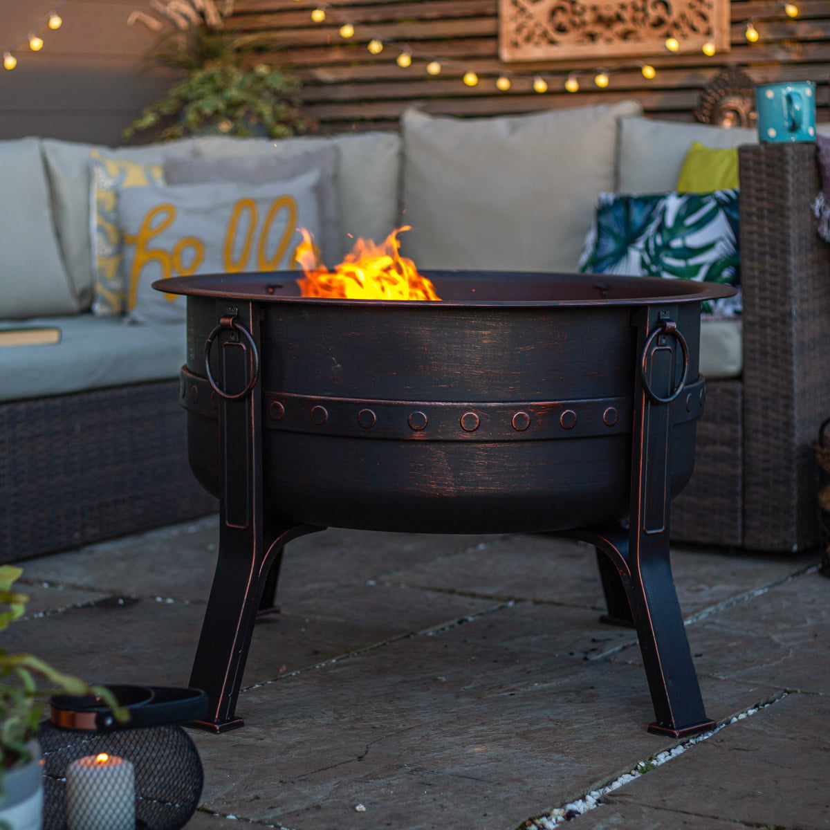 A La Hacienda Brava Firepit, designed with a black round structure and bronze effect, rests on a stone patio, its flames flickering warmly. It's encircled by outdoor furniture adorned with cushions and decorative pillows. Steel string lights dangle overhead, casting a cozy glow as plants rustle softly in the foreground.