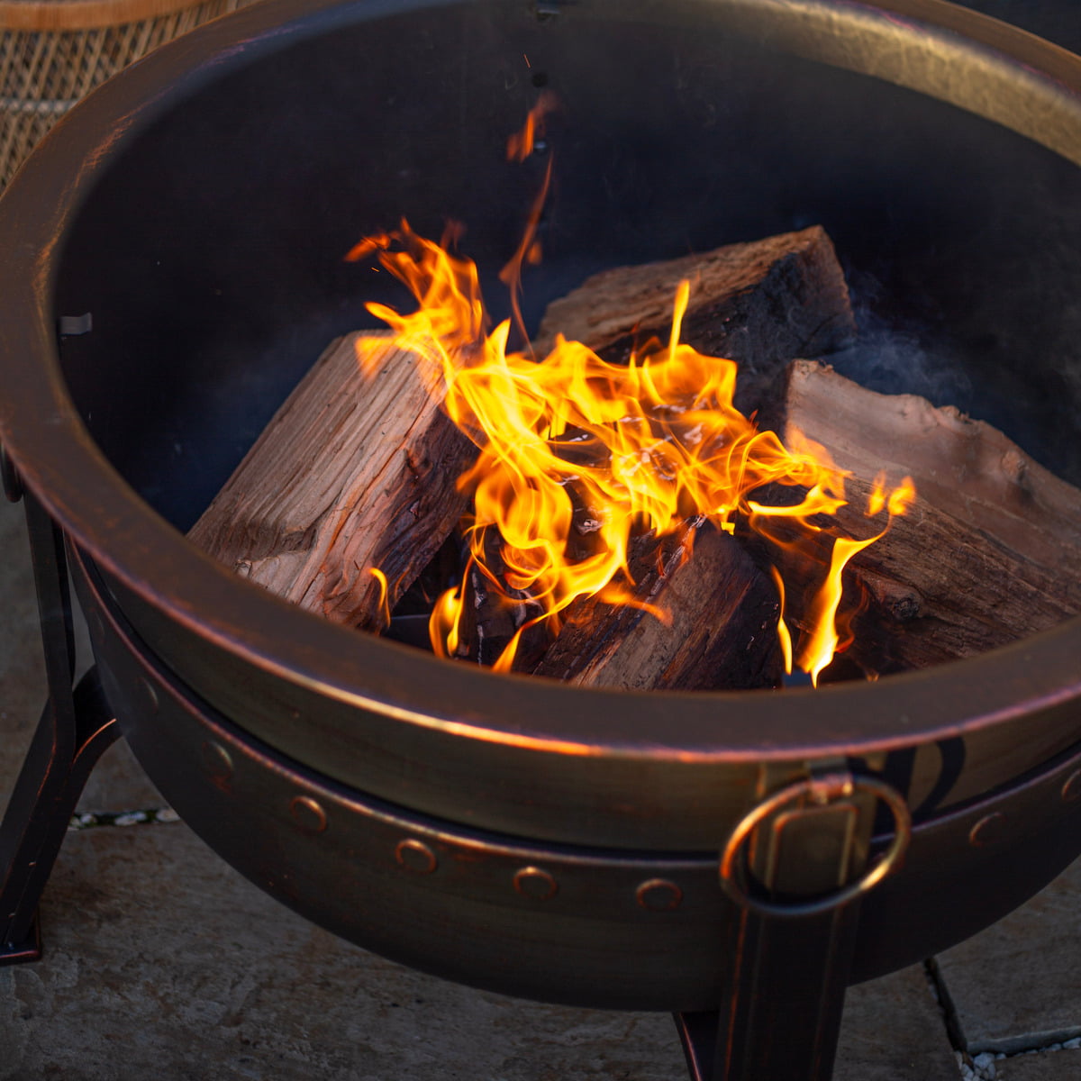 A close-up of the La Hacienda Brava Firepit reveals bright orange flames dancing over the burning logs, casting a warm glow. The bronze effect on the steel of this La Hacienda firepit enhances its charm as it rests on a stone surface, creating a cozy outdoor ambiance.