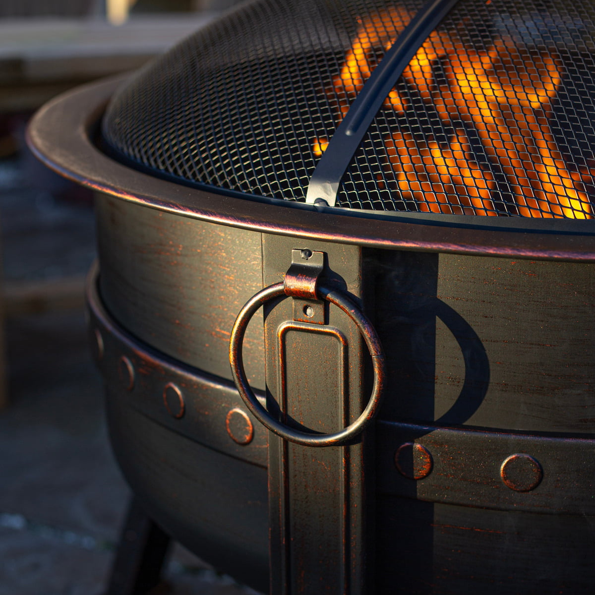 A close-up of the La Hacienda Brava Firepit reveals a warm, glowing fire within its mesh-covered, bronze effect design. A sturdy handle is attached to the side, and its steel surface features a rustic, weathered texture.