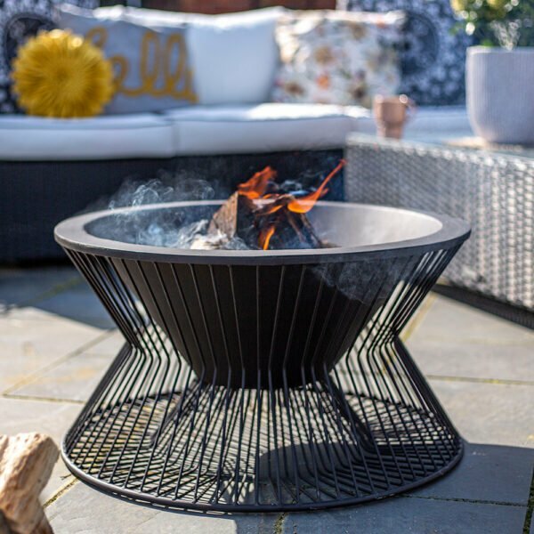 The La Hacienda Milan Firepit, featuring a modern design and robust steel construction, presents vibrant orange flames against its elegant black frame. Set on a stone patio, it's surrounded by outdoor seating embellished with both patterned and solid-colored cushions, as smoke softly drifts into the evening sky.