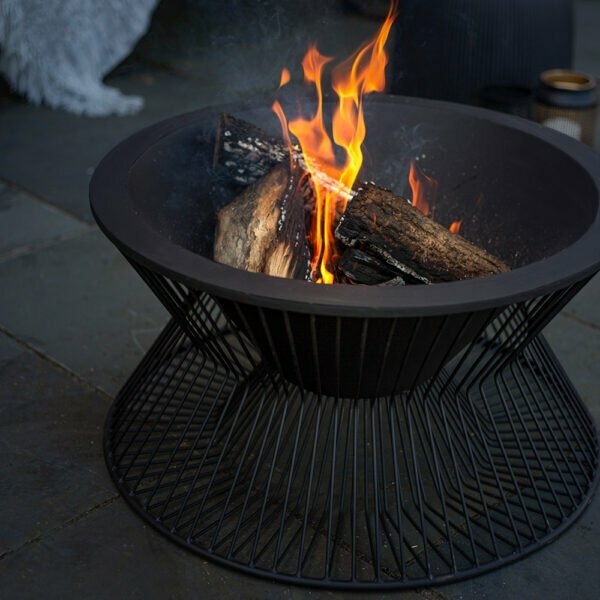 A modern fire pit with a sleek wireframe base on a patio features the contemporary design of the La Hacienda Milan Firepit. Burning logs with bright orange flames create a cozy ambiance, enhanced by the warm glow and nearby soft furnishings. Its durable steel construction ensures lasting enjoyment.