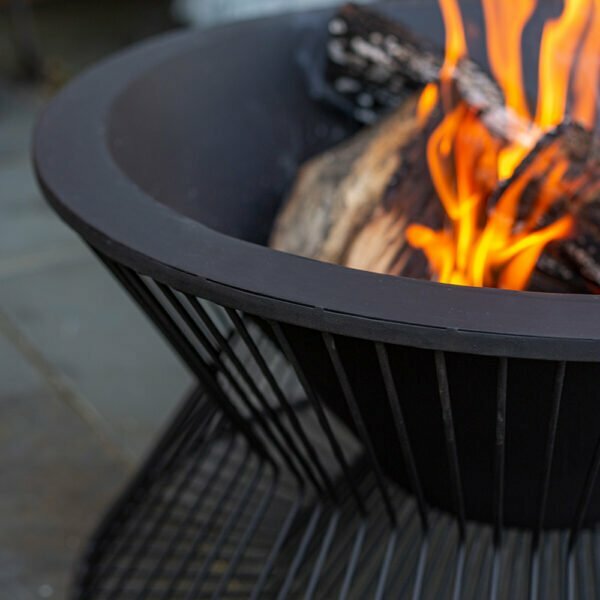 A detailed look at the La Hacienda Milan Firepit showcases its striking contemporary design, featuring vibrant orange and yellow flames flickering above burning wood. Constructed with durable steel, this sleek black fire pit from La Hacienda is set against a backdrop of gray stone tiles.