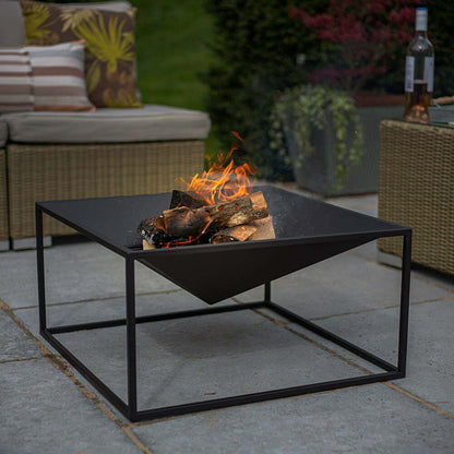 The La Hacienda Cairo Firepit features a modern design with sleek geometric lines, perfectly burning wood on the patio. It is surrounded by wicker furniture adorned with patterned cushions, while nearby plants enhance the natural feel of the setting. A bottle of wine and a glass sit elegantly on a table in the background, completing the sophisticated atmosphere.