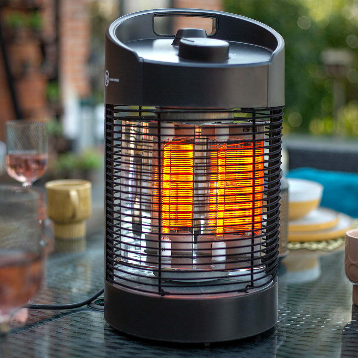 The La Hacienda Tabletop Heater, a cylindrical electric device featuring a black metal grill, emits a warm glow amidst the blurred tableware on the glass surface. Designed to be eco-friendly and suitable for IPX4 outdoor use, it enhances the lush greenery and brick wall backdrop of the patio setting.