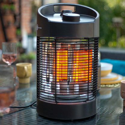 The La Hacienda Tabletop Heater, a cylindrical electric device featuring a black metal grill, emits a warm glow amidst the blurred tableware on the glass surface. Designed to be eco-friendly and suitable for IPX4 outdoor use, it enhances the lush greenery and brick wall backdrop of the patio setting.