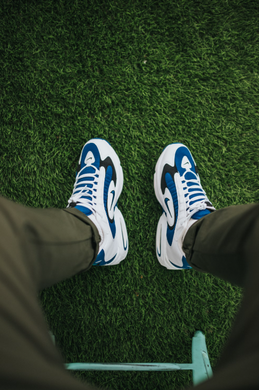 A person in blue and white sneakers with a wave pattern sits on Deck&Grass's Premium Artificial Grass, enjoying its lush 30mm-50mm blades. Their olive green pants blend seamlessly with the low-maintenance, UV-resistant grass, keeping the scene vibrant and inviting.