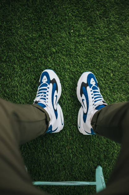 A person in blue and white sneakers with a wave pattern sits on Deck&Grass's Premium Artificial Grass, enjoying its lush 30mm-50mm blades. Their olive green pants blend seamlessly with the low-maintenance, UV-resistant grass, keeping the scene vibrant and inviting.