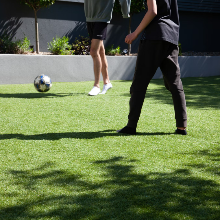 Two people enjoy a game with a soccer ball on Deck&Grass Budget Artificial Grass. They are casually dressed in shorts and pants, with one wearing a hoodie. The sunny, outdoor scene is complete with shadows and a garden visible in the background.