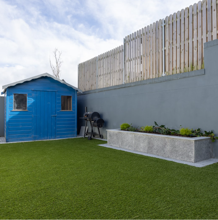 A blue wooden shed sits on Deck&Grass Budget Artificial Grass, accompanied by a black barbecue grill. A gray concrete planter with small plants lines the right wall, topped with a wooden fence under a partly cloudy sky.