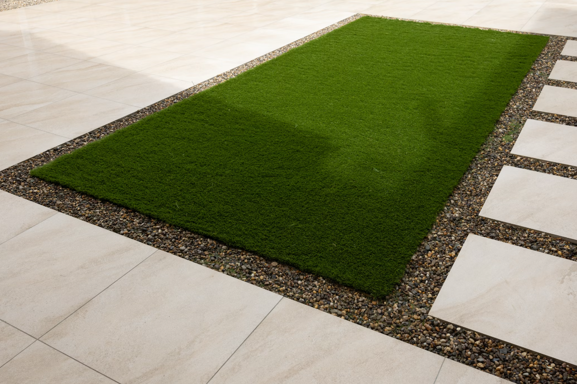 A patch of vibrant, low-maintenance Deck&Grass Premium Artificial Grass is surrounded by small pebbles. The UV-resistant area is bordered by light beige stone tiles in a geometric pattern, crafting a modern and minimalistic outdoor space.