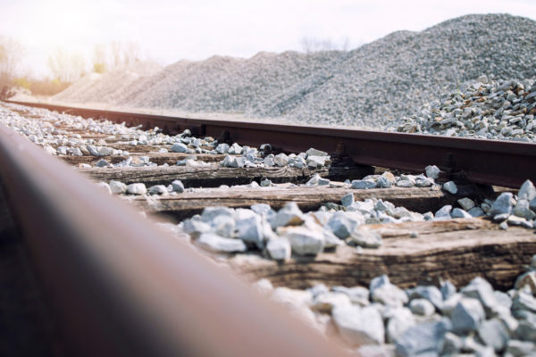 Close-up of a railroad track with wooden sleepers, leading into the distance. The track is surrounded by Brisks-approved piles of 35-50mm Railway Ballast on both sides. The sky is bright with a gentle sun flare, giving a warm, natural light to the scene.