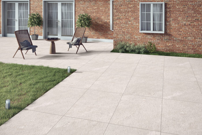 A patio area featuring light gray, slip-resistant tiles that are ideal for outdoor spaces. Two wicker chairs sit alongside a small table on Brisks' Agra White Porcelain Paving slabs. The scene is set against a red brick wall with double glass doors and windows, while potted plants and patches of grass provide additional greenery around the patio.