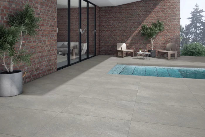 Brisks' Ancient Grey Porcelain Paving tiles adorn the modern outdoor patio, elevating its contemporary design. A swimming pool lies in the foreground, accompanied by a seating area adjacent to a glass wall and brick exterior. Potted plants contribute greenery, while trees provide a scenic backdrop.