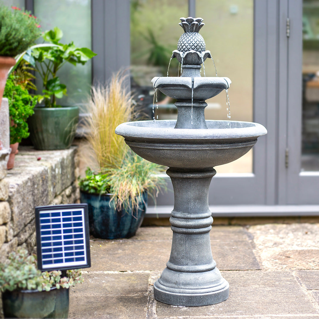 A gray Angelica Tiered Fountain by La Hacienda, featuring two tiers and a charming pineapple design on top, graces the patio near potted plants. Its solar panel discreetly hints at its solar-powered water feature capability. Glass doors provide a backdrop to this antique stone design marvel.