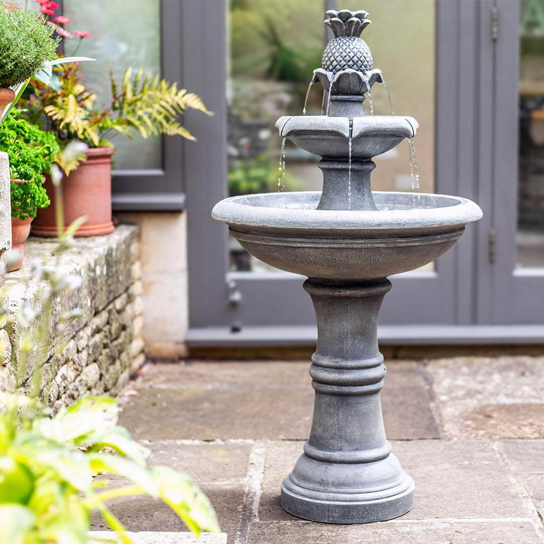 The Angelica Tiered Fountain by La Hacienda, showcasing an antique stone design topped with a decorative pineapple, is encircled by flowing water on a stone patio. Nearby, potted plants and ferns flourish as a glass door shimmers in the background.