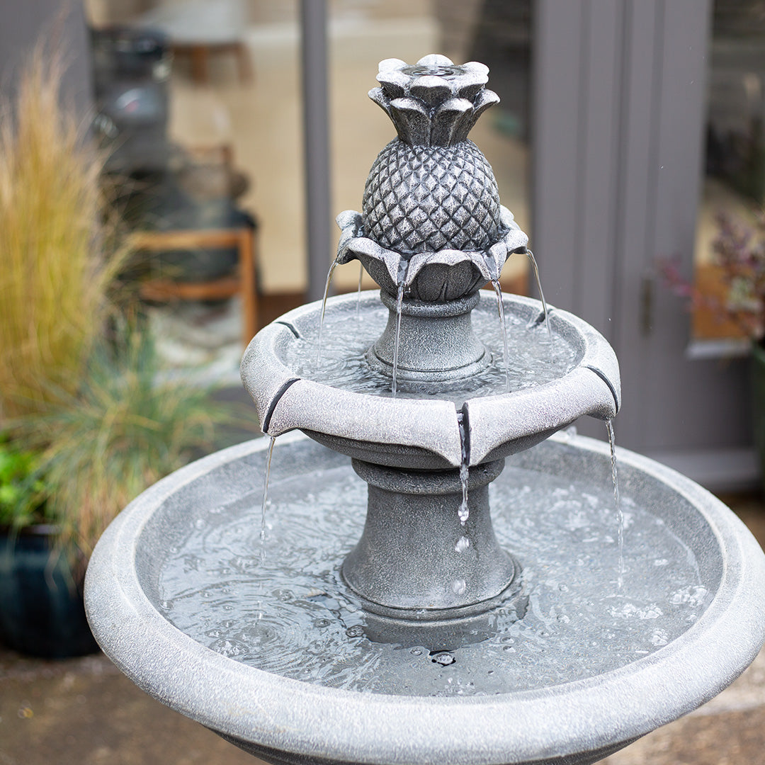 The La Hacienda Angelica Tiered Fountain, featuring an antique stone design and a pineapple motif at the top, is a charming addition. Water cascades gracefully from this solar-powered water feature into its tiered basin, set against a backdrop of glass doors and lush potted plants.