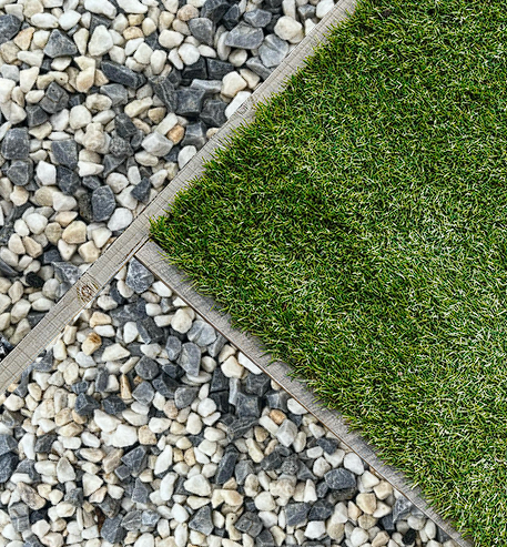 A close-up view of a garden featuring a border of wooden planks separating a section of artificial green grass on the right from Brisks 10-20mm Arctic Blue Chippings on the left. The clean lines create a contrast between the natural and synthetic materials.