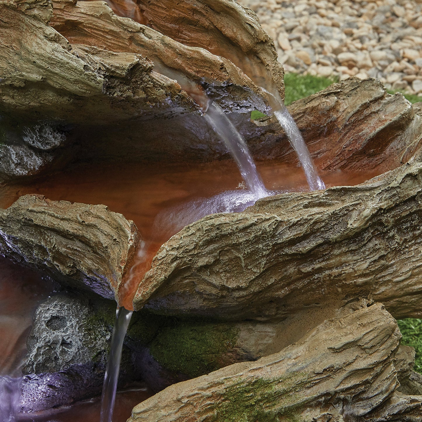 A rustic, multi-tiered La Hacienda Bubbling Brook (Inc. LED) Water Feature from La Hacienda, crafted from high-quality resin, features several small streams of water cascading down its contoured, bark-like surfaces into shallow pools. Enhanced with LED lights, the feature is set against a background of grass and pebbled ground.