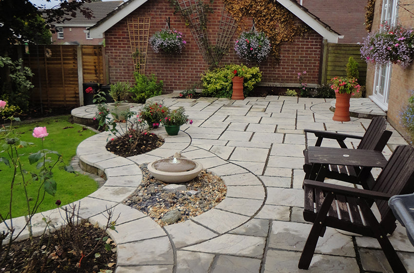 A beautifully landscaped backyard features a large, neatly laid stone patio with curved edges crafted from the premium Weathered Moss Cathedral Paving Kit by Brisks. The patio includes flower beds, a small fountain, hanging flower baskets, and several decorative plants. Wooden chairs and a table are positioned on the patio.