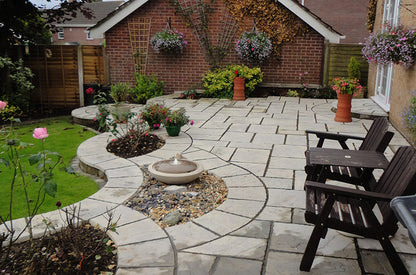 A beautifully landscaped backyard features a large, neatly laid stone patio with curved edges crafted from the premium Weathered Moss Cathedral Paving Kit by Brisks. The patio includes flower beds, a small fountain, hanging flower baskets, and several decorative plants. Wooden chairs and a table are positioned on the patio.