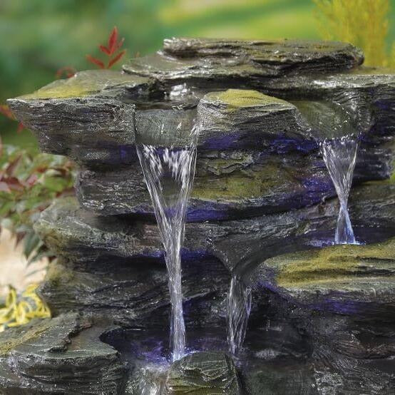 The La Hacienda Como Springs (Inc. LED) Water Feature features a durable resin structure and a small artificial waterfall composed of stacked dark rocks. Clear water flows gently over the rocks, creating multiple small cascades. LED lights illuminate the scene, while foliage and green moss enhance its natural appearance.