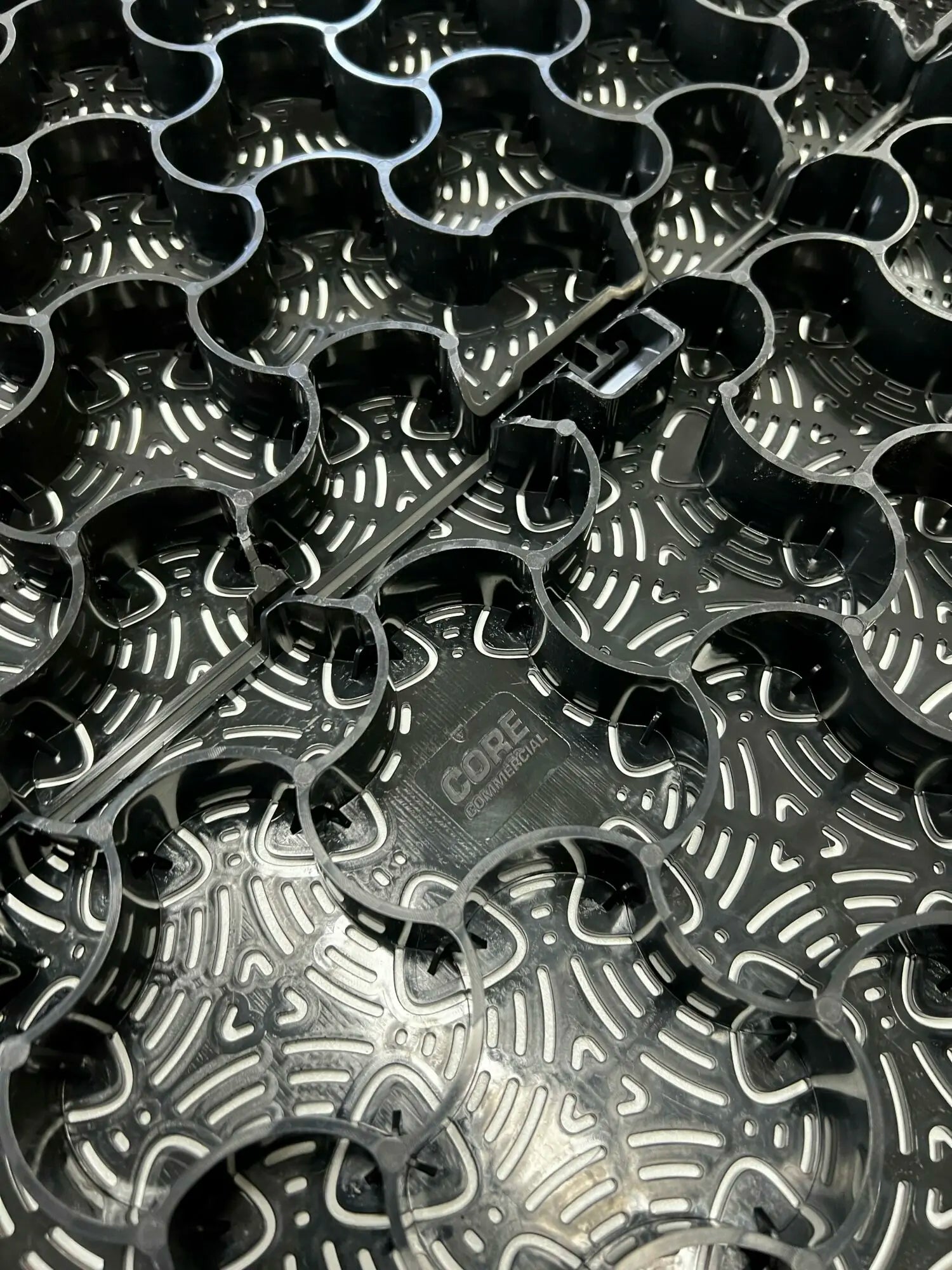 Close-up of a black plastic gravel stabilization mat named CORE COMMERCIAL 75-30 by Core LP, showcasing interlocking circular patterns with "CORE" at the center. The shiny surface features curved lines and notches.