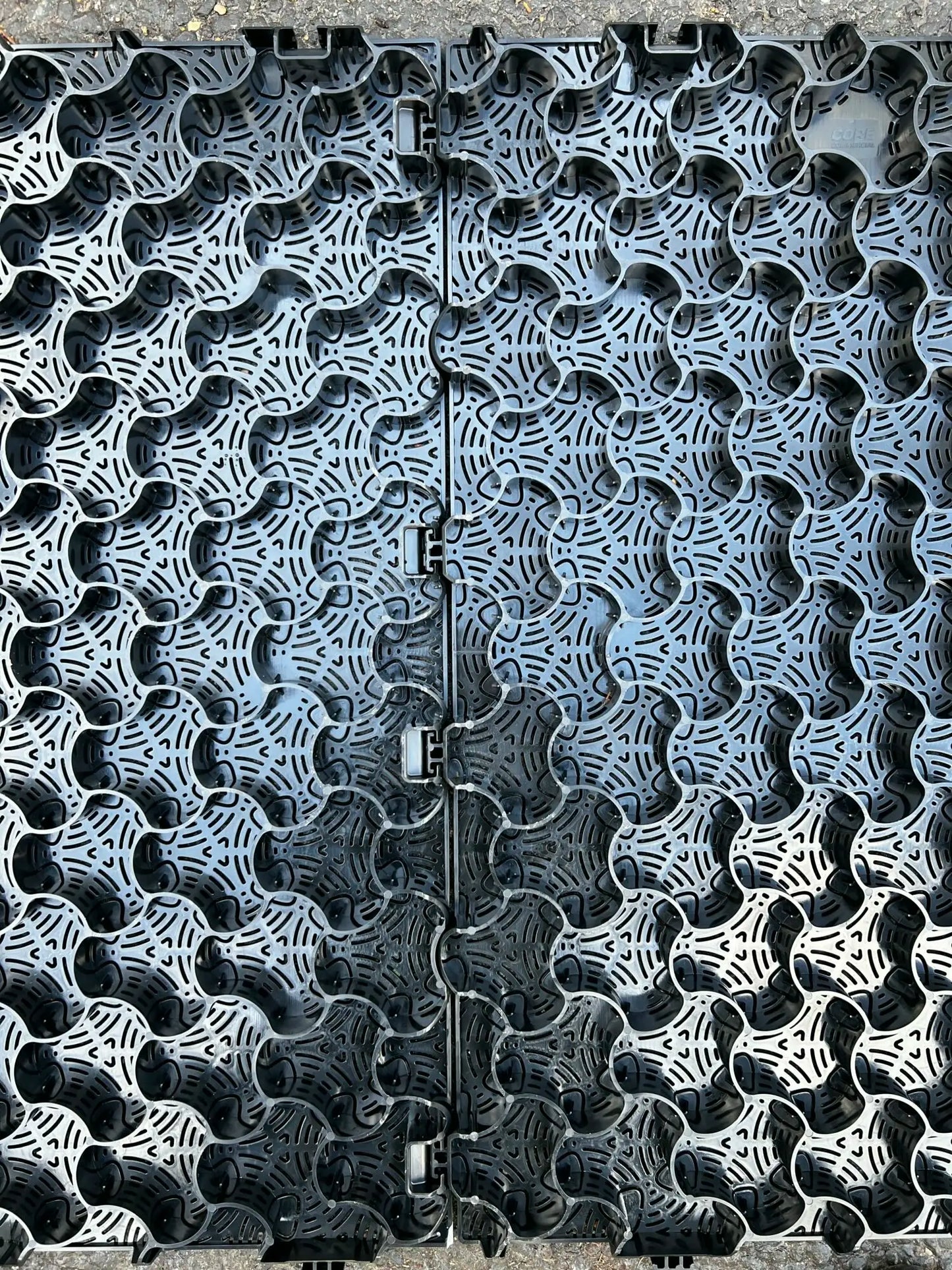 Close-up of Core LP's CORE COMMERCIAL 75-30 - Gravel Grid, made of interlocking black plastic with circular and curved shapes for gravel stabilization. The grid features center connectors, forming a textured, perforated surface suitable for managing commercial traffic.