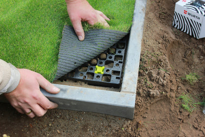 Hands delicately position a piece of artificial grass on a CORE DRAINBASE by Core LP, made from recycled HDPE, next to a concrete border. A box emblazoned with the brand logo is visible in the background.