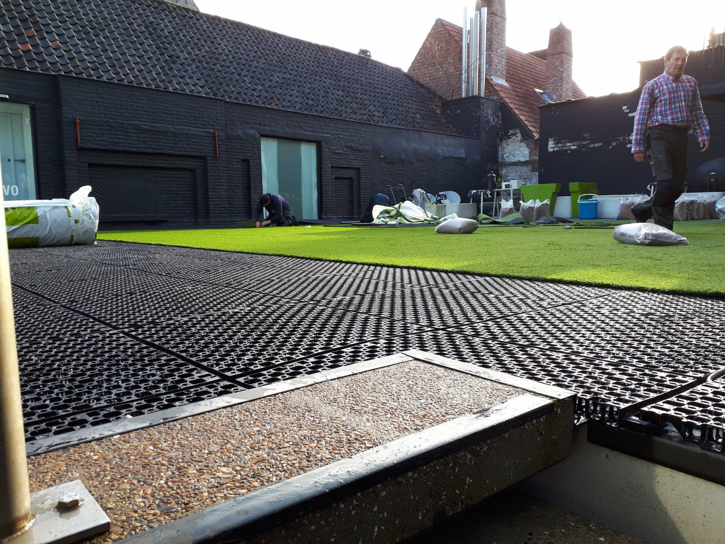 The rooftop garden installation features CORE DRAINBASE tiles from Core LP, with black grids and patches of artificial grass for efficient drainage. Two workers are present—one walking and one kneeling—as buildings with sloped roofs rise in the bright sky background.