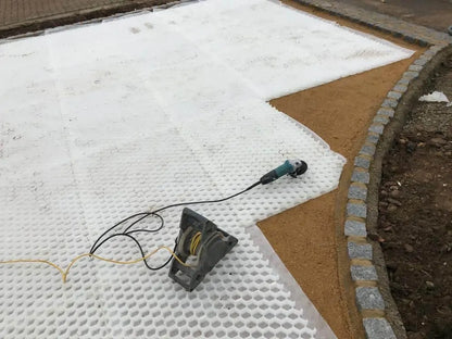 The construction site features a CORE DRIVE 50-35 gravel grid from Core LP for gravel stabilization. A power tool with cables is on the surface, while brown gravel lines the edges, bordered by a curved line of gray cobblestones creating an eco-friendly driveway.