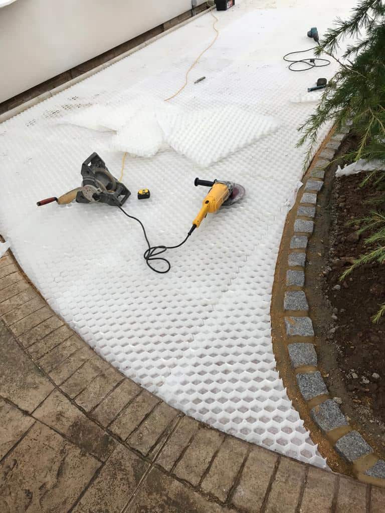 On a curved outdoor pathway under construction, the CORE DRIVE 50-35 - Gravel Grid by Core LP, made from recyclable HDPE, is laid out. A power tool and tape measure are on the mat to help complete this eco-friendly driveway with stone edging on one side and soil on the other.