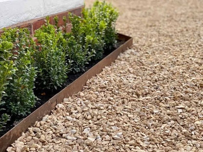 A garden with a neat border of green shrubs next to a brick wall is enhanced by Core LP's durable CORE EDGE lawn edging. The ground is covered in light brown gravel, creating a tidy and organized appearance.