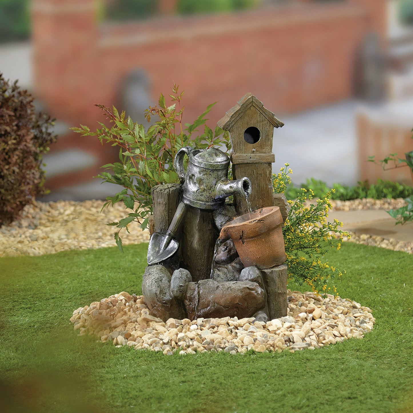 Introducing the La Hacienda Country Garden Pour (inc LED) Water Feature by La Hacienda: a delightful garden decoration showcasing a poly-resin birdhouse perched atop a wooden post. This charming scene includes a watering can, a small shovel, a terracotta pot, and an endearing figurine of a dog resting peacefully. All these elements are artistically arranged on a circular bed of white pebbles, with grass and plants providing a picturesque backdrop.