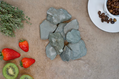 A flat lay image shows Brisks 20mm Green Slate Chippings in the center, fresh thyme on the upper left, strawberries and sliced kiwi at the bottom left, and a white cup with coffee beans on a saucer at the upper right, all arranged on a beige textured surface reminiscent of garden borders.