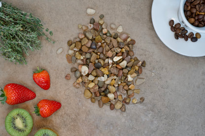 A collection of various small stones and Brisks 04-10mm Pea Gravel is spread on a beige surface. Surrounding the stones are a sprig of green herbs, three strawberries, a sliced kiwi, and a white cup with coffee beans on a saucer. The arrangement suggests a natural and earthy theme perfect for landscaping inspiration.
