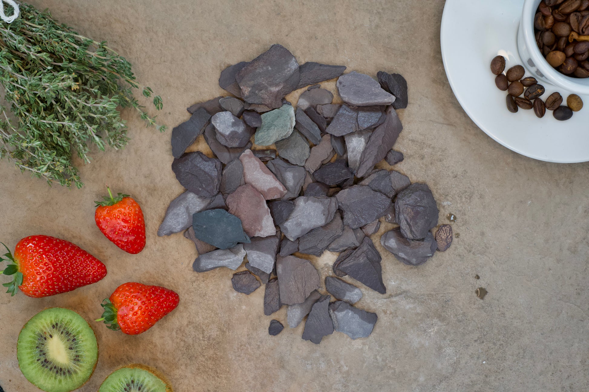 A display of various items: a cluster of fresh thyme, a few ripe strawberries, sliced kiwis, a cluster of Brisks 20mm Plum Slate Chippings, and a white plate with coffee beans. All items are arranged on a textured, beige surface.