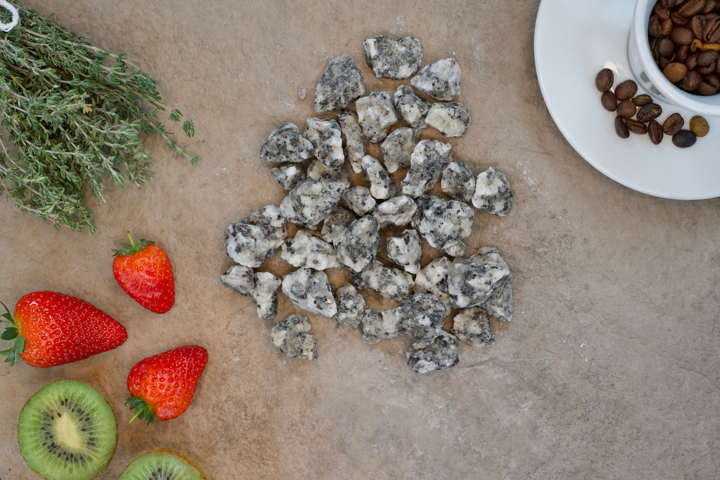 A collection of small, gray, rocky stones, resembling Brisks 20mm Black and White Granite Chippings, are centrally placed on a beige surface. Surrounding the stones are a sprig of thyme, a few strawberries, a slice of kiwi, and a white saucer containing coffee beans. The scene seems to depict various natural elements.