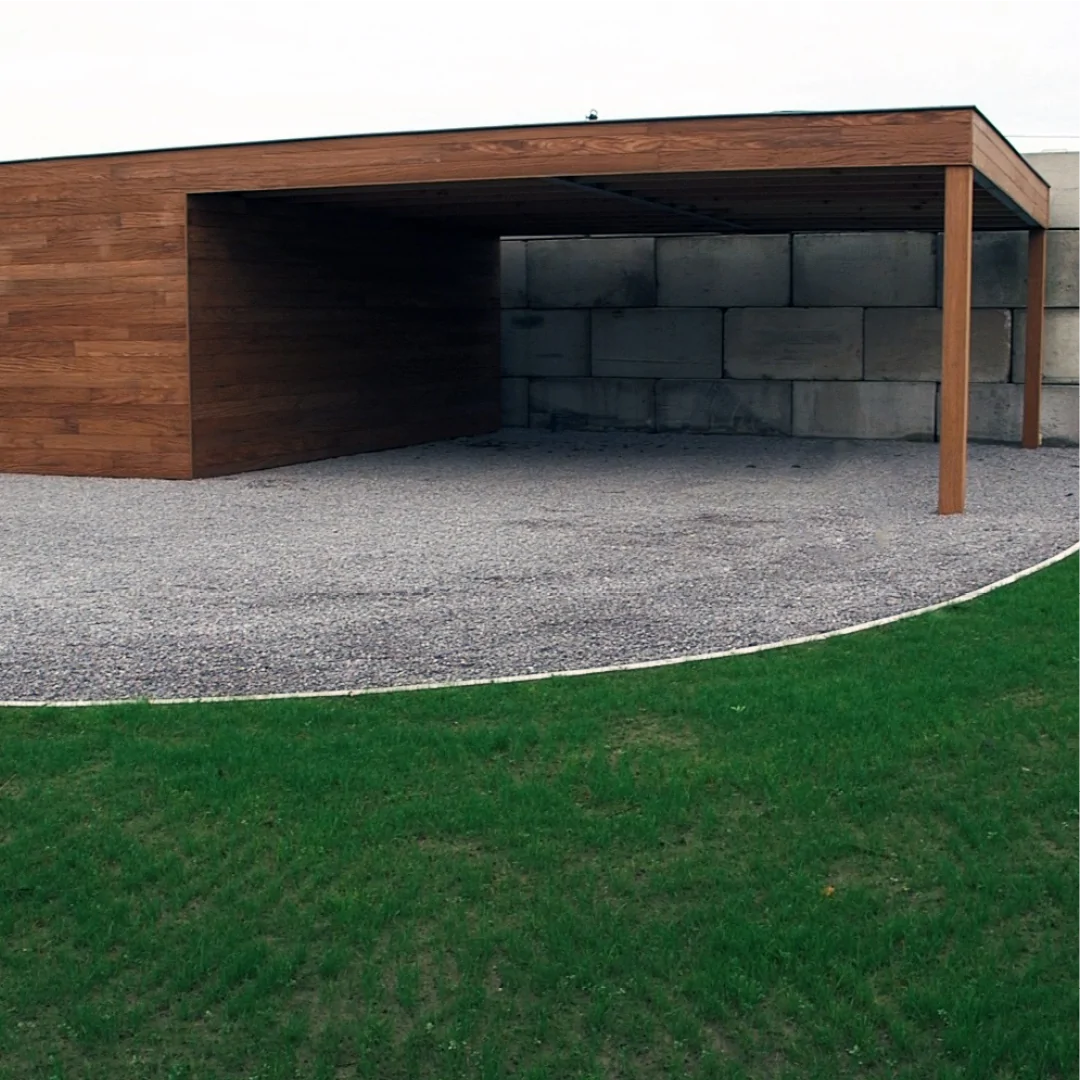 A modern wooden carport with a flat roof and open sides sits on a gravel surface next to a lush grassy area, backed by a tall stone wall. Designed for minimalism and sleekness, it incorporates the CORE GRASS 40 grass reinforcement grid by Core LP, measuring 800 x 800 mm (0.64 m²), providing added stability and eco-friendliness for light commercial traffic.