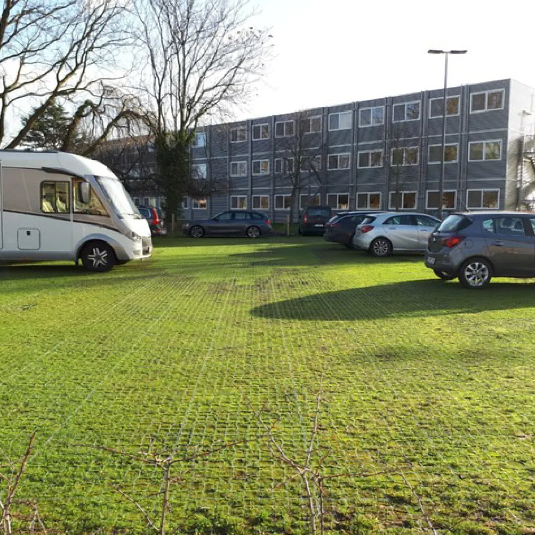 Several cars and a white camper van are parked on a grassy area next to a multi-story building. The ground, reinforced with CORE GRASS 40 by Core LP, made from recycled high-density polyethylene (HDPE), facilitates drainage. Bare trees are visible in the background under a clear sky.