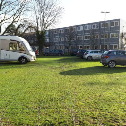 Several cars and a white camper van are parked on a grassy area next to a multi-story building. The ground, reinforced with CORE GRASS 40 by Core LP, made from recycled high-density polyethylene (HDPE), facilitates drainage. Bare trees are visible in the background under a clear sky.