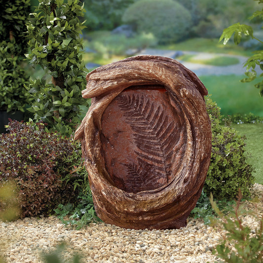 A decorative garden feature from La Hacienda, the Forest Fern (Inc. LED) Water Feature, showcases a rustic wooden frame that encloses a textured, rust-colored panel with a fern imprint. Surrounded by small pebbles, lush green bushes, and a vibrant garden in the background, the scene is enhanced by soft LED lights.